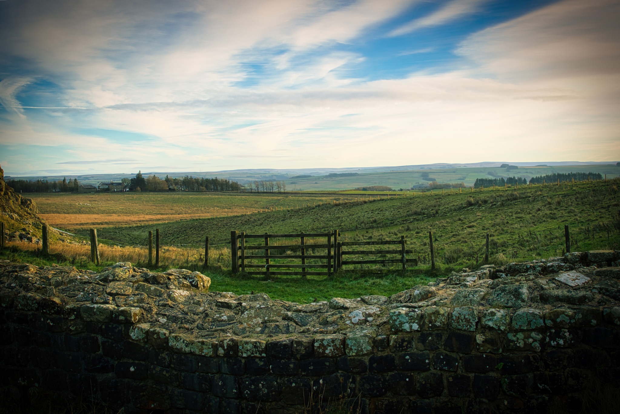 Rustic wooden gate on a stone wall with expansive countryside views under a vast sky at Sycamore Gap, Northumberland, UK.