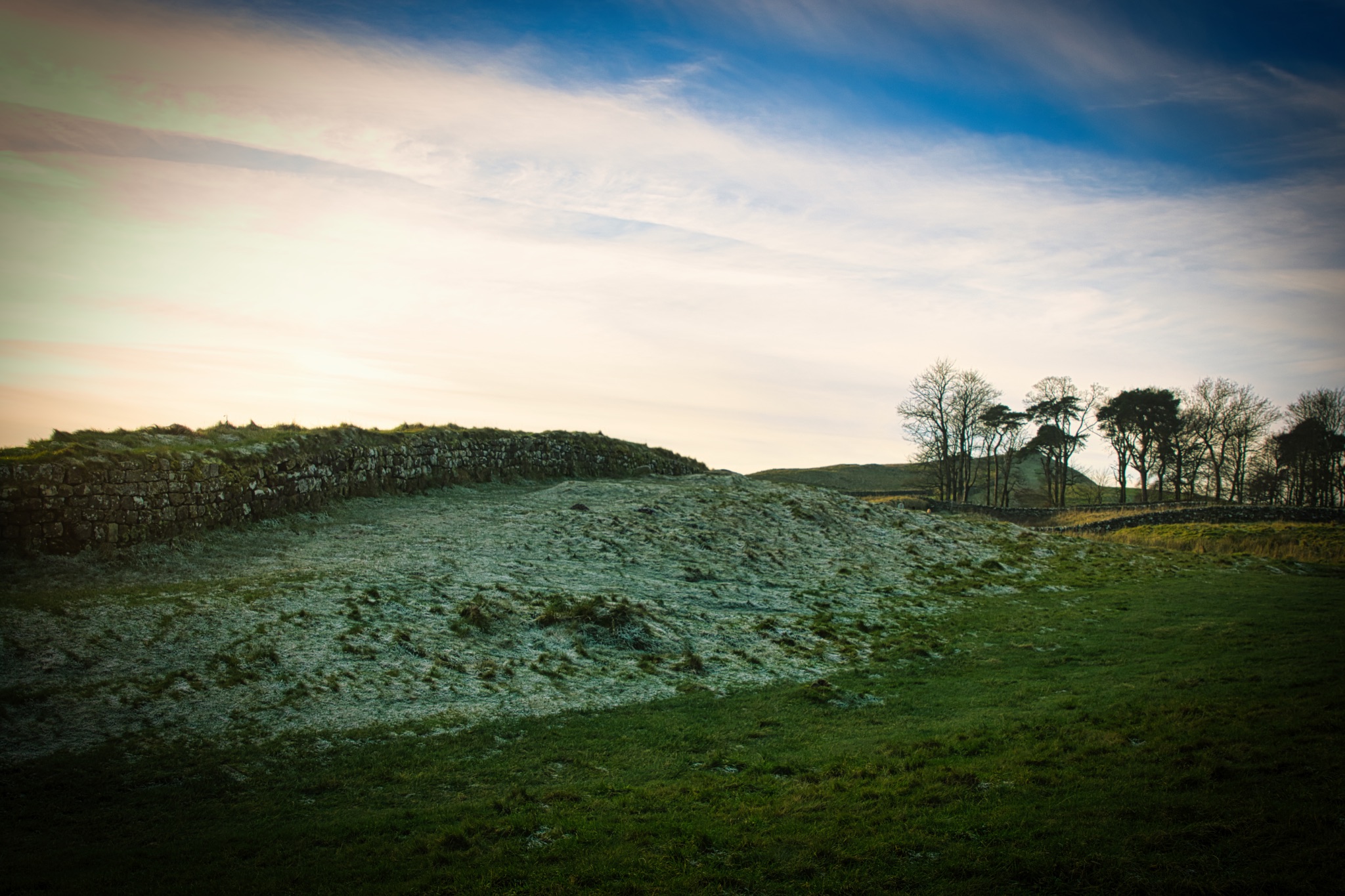 Serene landscape at dusk with vibrant sky, stone wall, and silhouetted trees on a hill at Sycamore Gap, Northumberland, UK.