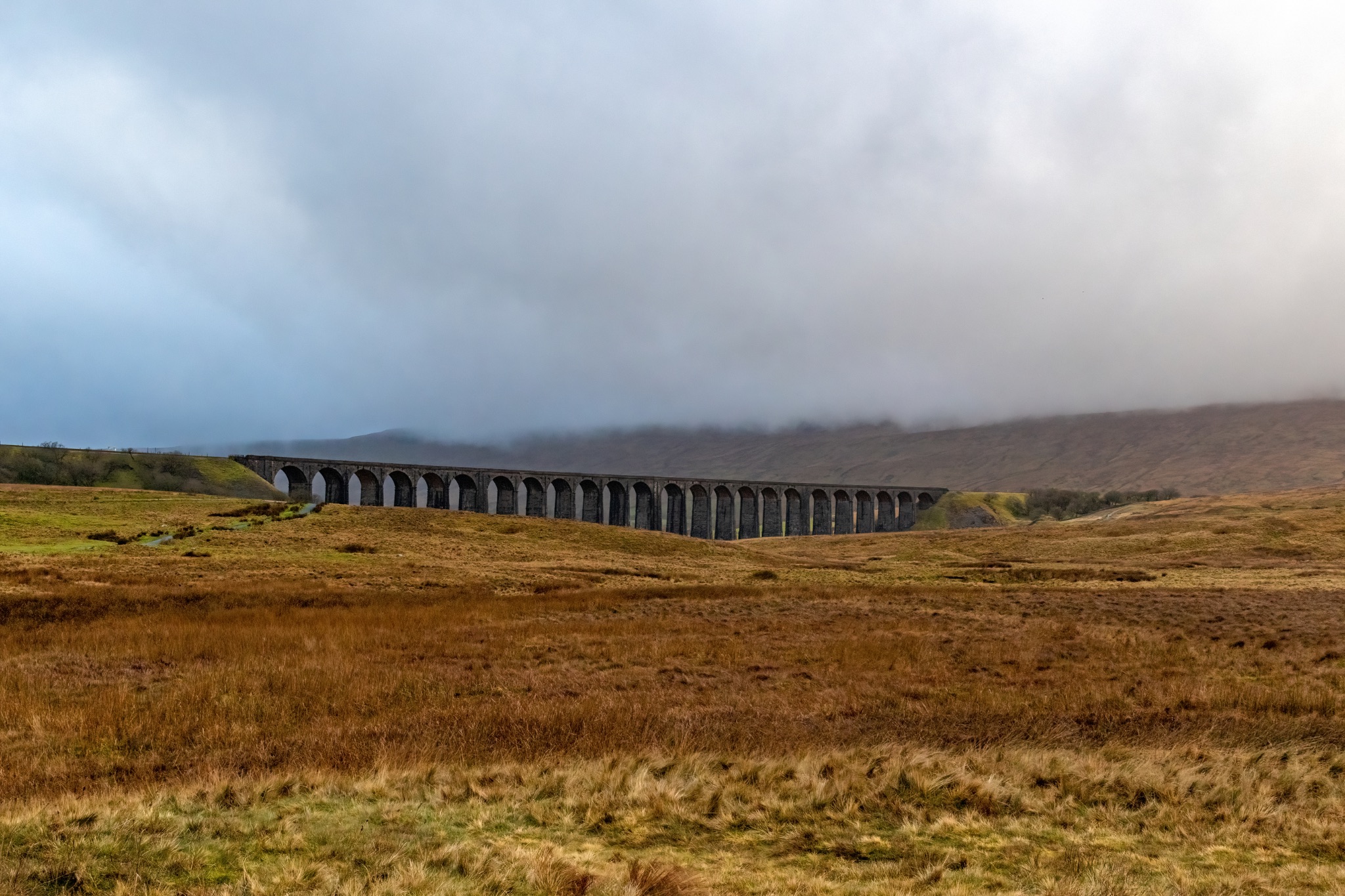 Scenic view of a historic viaduct in a misty landscape with rolling hills and overcast skies at Ribblehead Viaduct, Yorkshire.