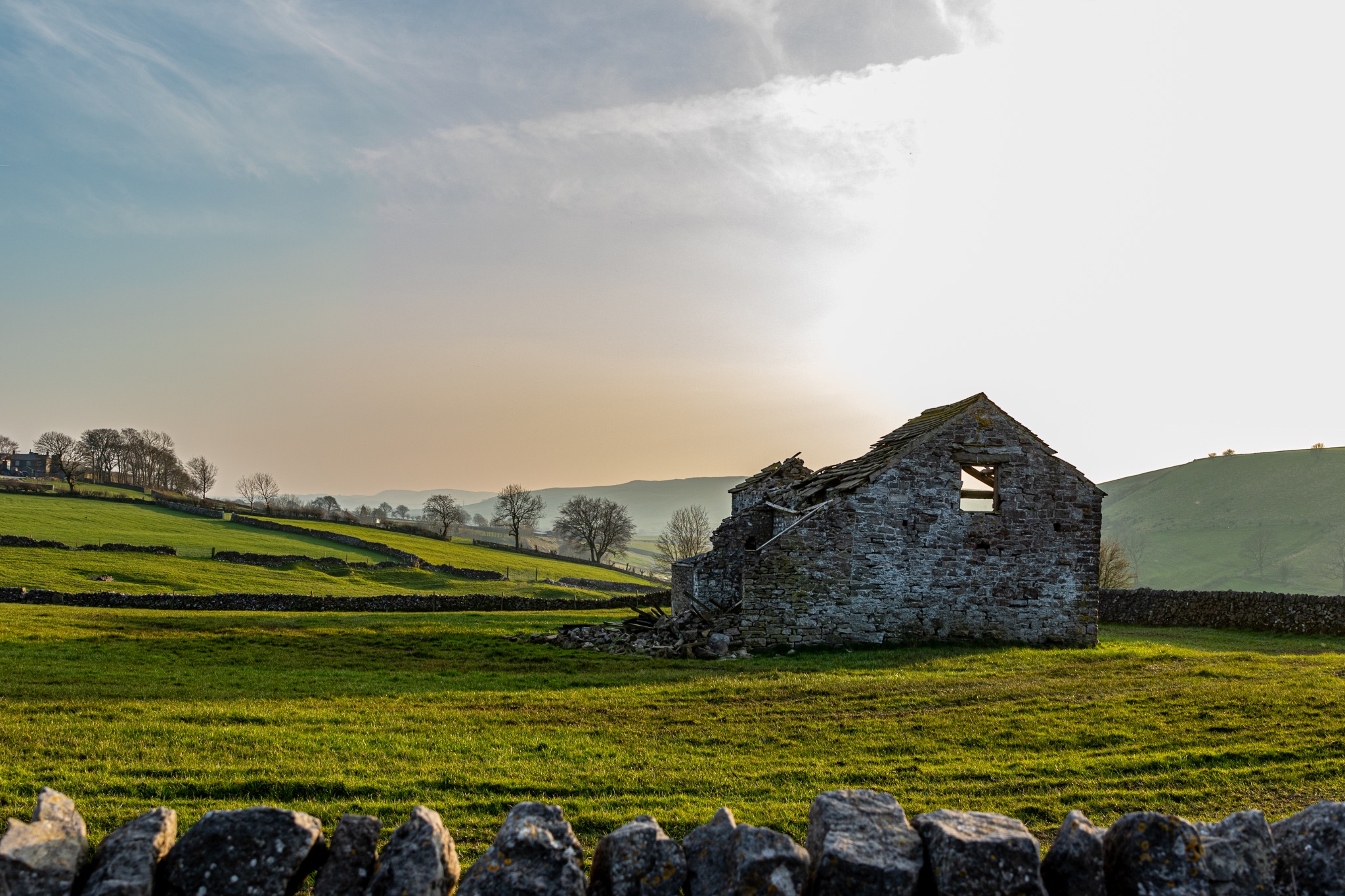 Ruined stone cottage in lush green countryside with a stone wall in the foreground and a sunset sky.