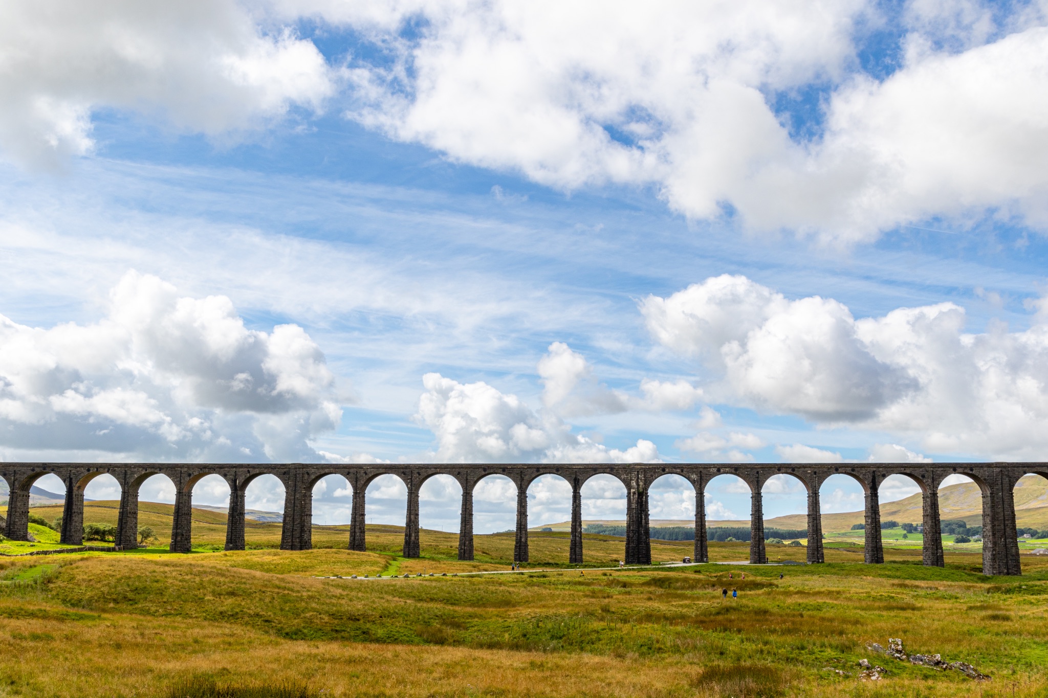 Scenic view of a historic viaduct against a backdrop of blue sky with fluffy clouds, surrounded by lush greenery at Ribblehead Viaduct, Yorkshire