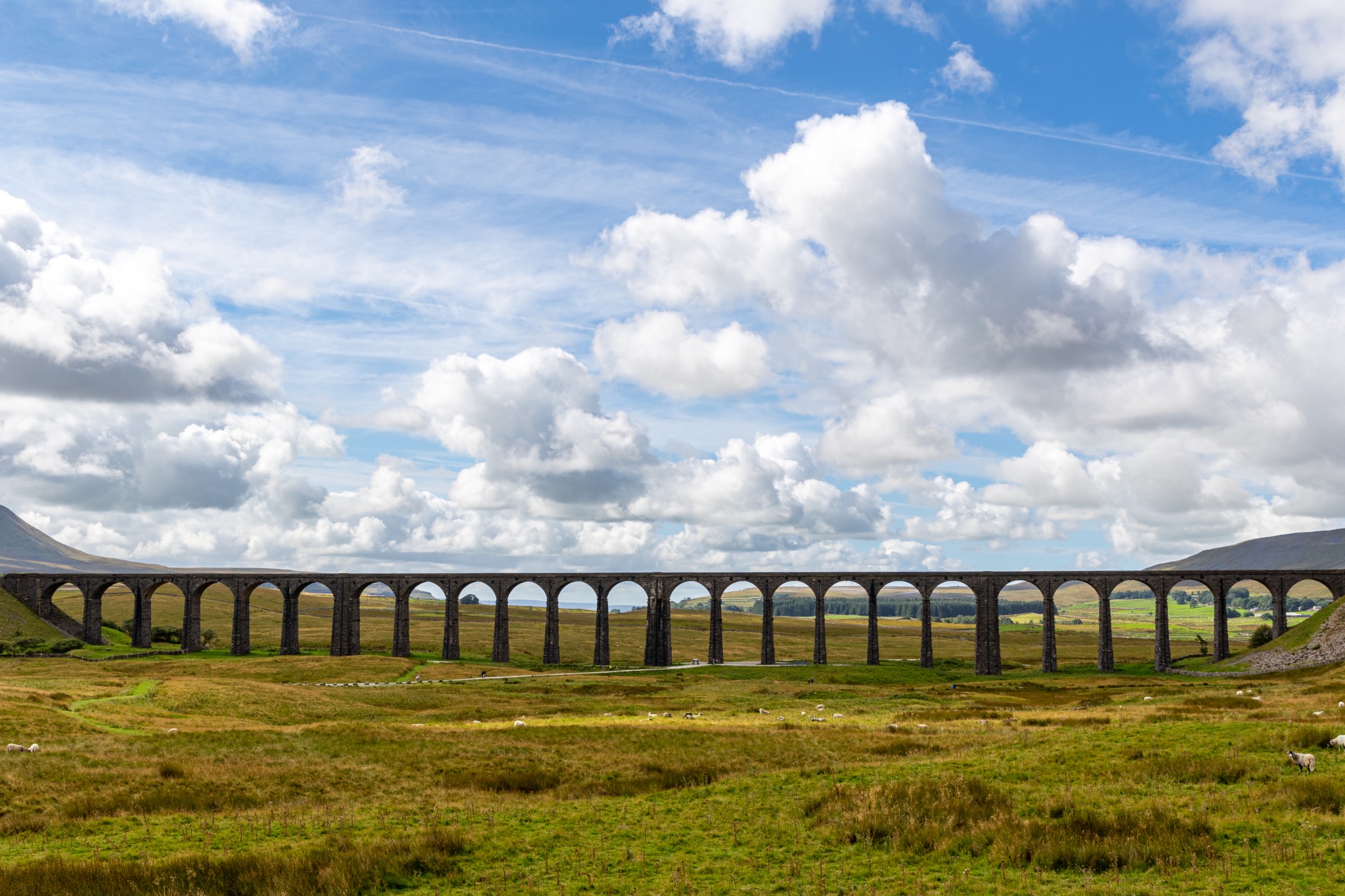 Scenic view of a historic viaduct in a lush green landscape under a cloudy blue sky at Ribblehead Viaduct, Yorkshire