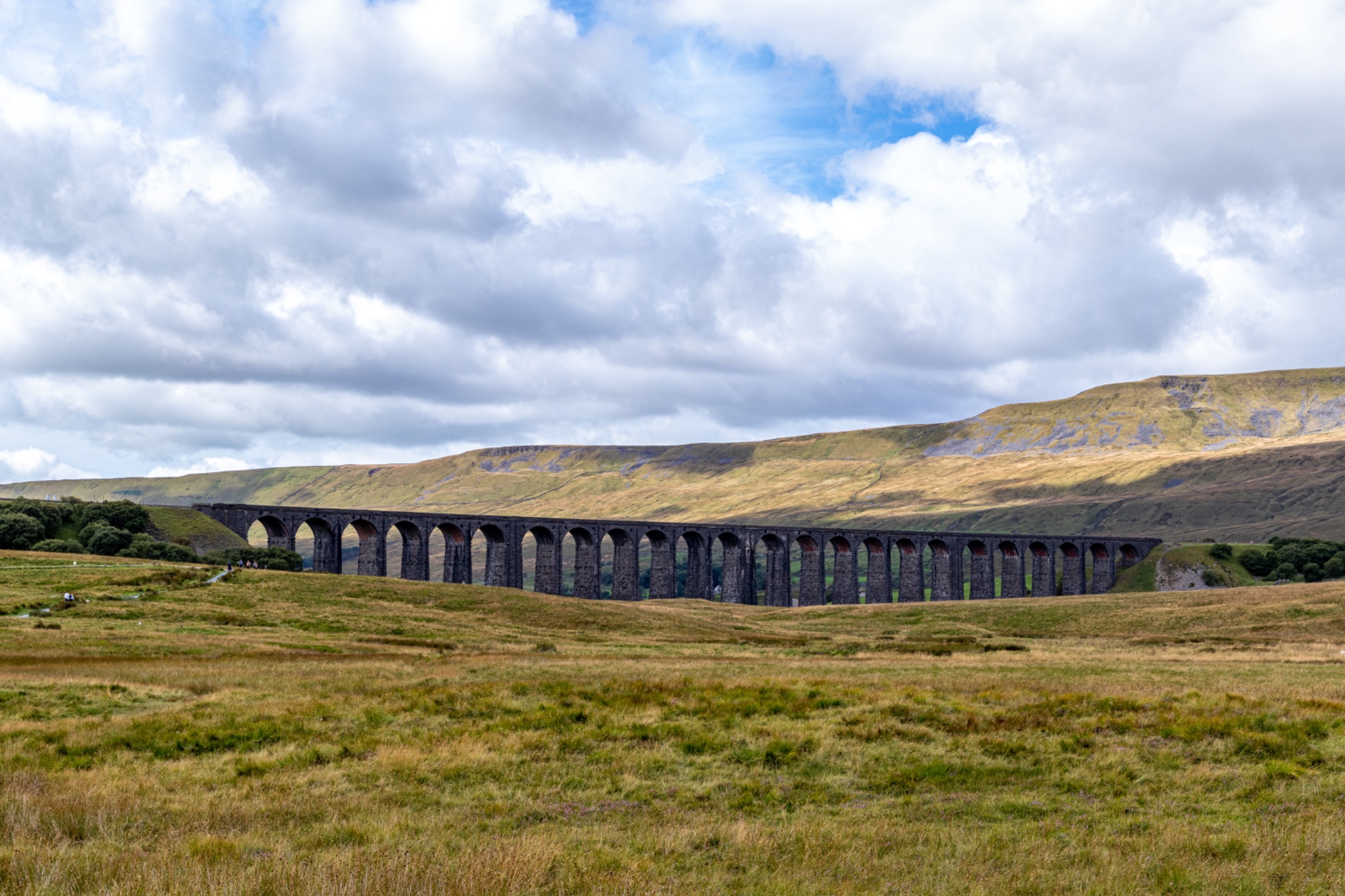 Scenic view of an old viaduct in a lush green valley with rolling hills and a cloudy sky in the background at Ribblehead Viaduct, Yorkshire